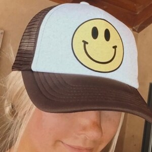May include: A brown and white trucker hat with a yellow smiley face on the front. The hat has a mesh back.