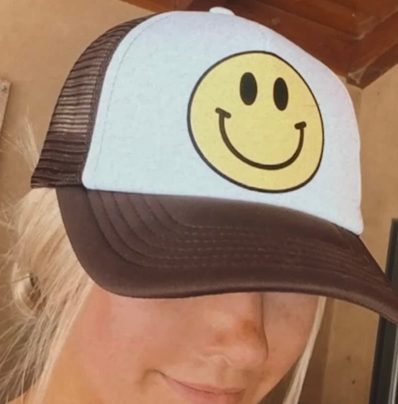 May include: A brown and white trucker hat with a yellow smiley face on the front. The hat has a mesh back.