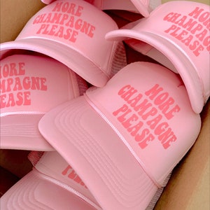 Op de afbeelding: Een stapel roze trucker caps met de tekst "More Champagne Please" in roze letters.