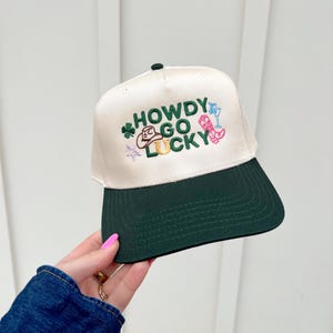 以下が含まれることがあります： 白いグリーンのベースボールキャップで、前面に「HOWDY GO LUCKY」という刺繍が施されています。キャップはグリーンのつばがあり、馬蹄、カウボーイハット、クローバー、ブーツなど、さまざまな刺繍されたアイコンが特徴です。