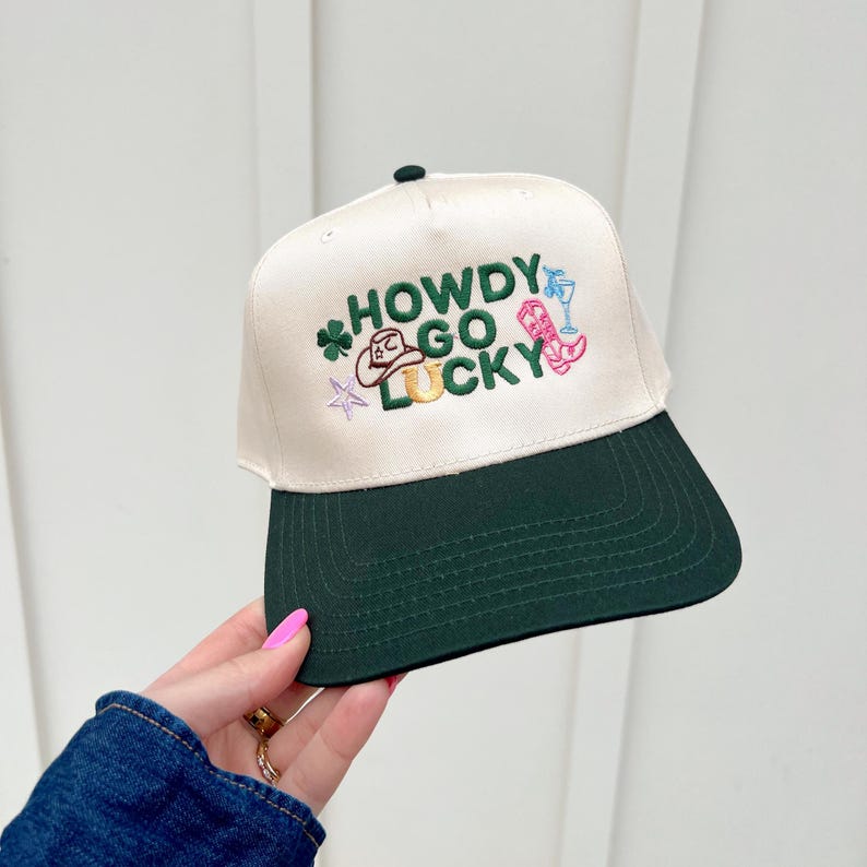 以下が含まれることがあります： 白いグリーンのベースボールキャップで、前面に「HOWDY GO LUCKY」という刺繍が施されています。キャップはグリーンのつばがあり、馬蹄、カウボーイハット、クローバー、ブーツなど、さまざまな刺繍されたアイコンが特徴です。