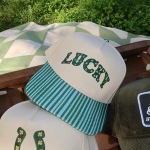 May include: A cream-coloured baseball cap with the word "LUCKY" embroidered in green and pink. The brim features green and blue stripes. Other caps are visible in the image, including one with a horseshoe design.