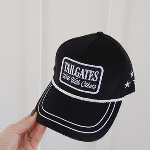 May include: A black corduroy baseball cap featuring a white rectangular patch with the text "TAILGATES Well With Others" in white embroidery. The cap includes a white braided rope trim across the base of the crown and white star embroidery.