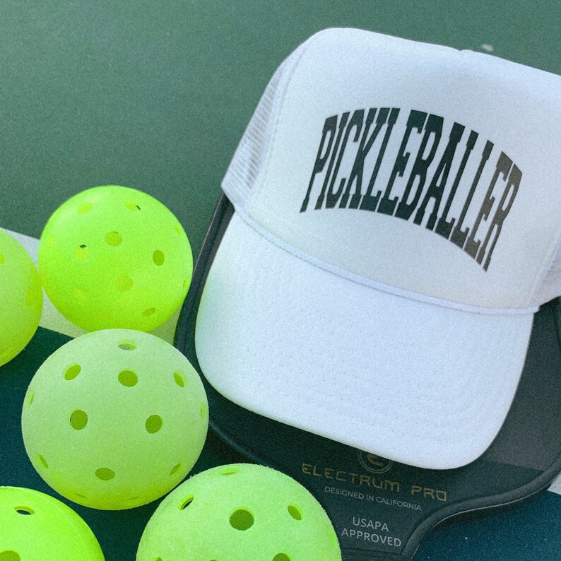 May include: A white mesh baseball cap with the text "Pickleballer" in black lettering. The cap is on top of a black and green pickleball paddle with four green pickleballs.