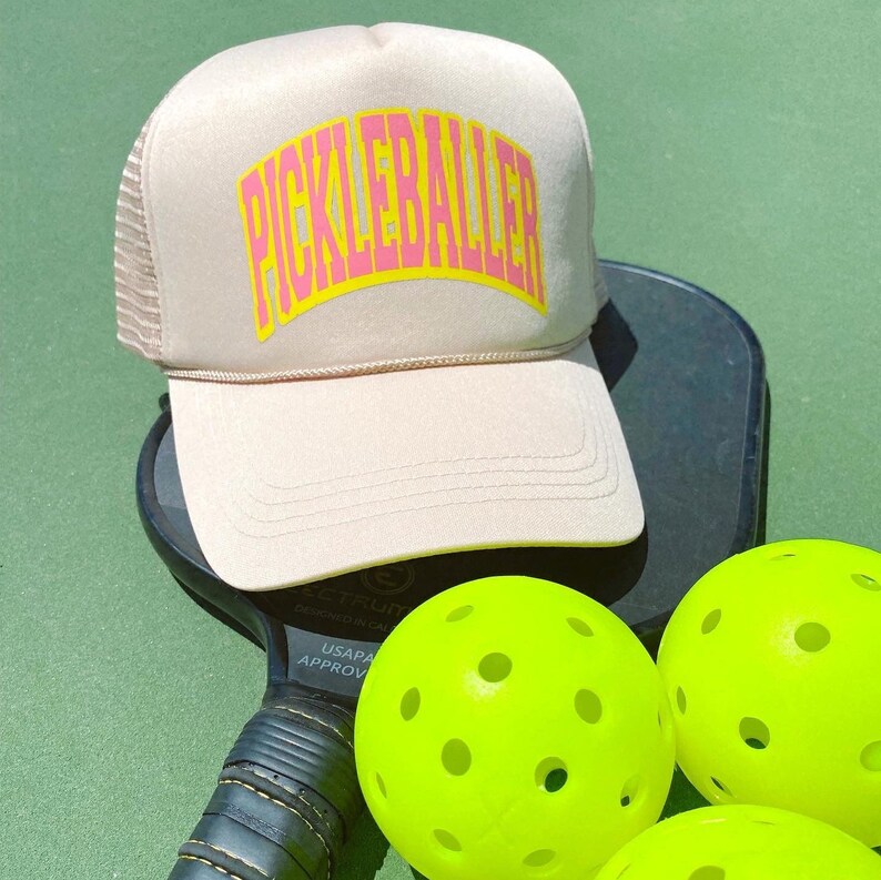 May include: A beige mesh trucker hat with a pink "Pickleballer" logo on the front. The hat is sitting on top of a black pickleball paddle with three yellow pickleballs in the foreground.