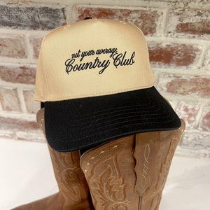 Op de afbeelding: Een beige baseballpet met een zwarte klep en de tekst "not your average Country Club" geborduurd in het zwart op de voorkant. De pet ligt op een paar bruine cowboylaarzen.