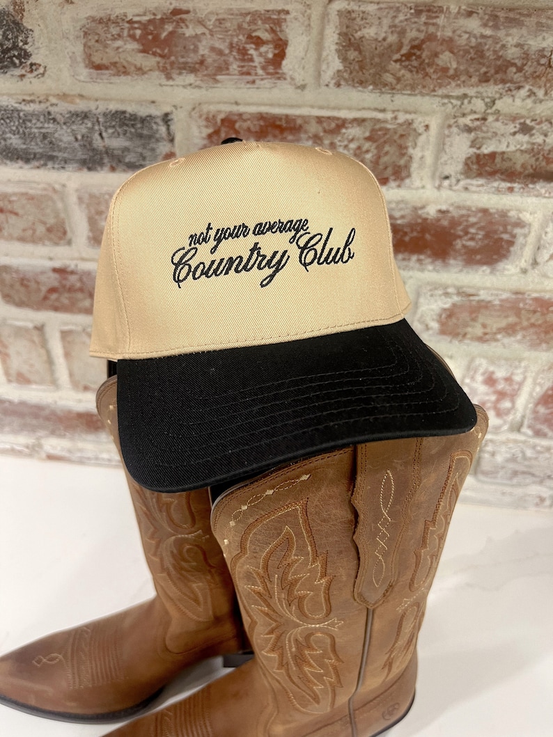 Op de afbeelding: Een beige baseballpet met een zwarte klep en de tekst "not your average Country Club" geborduurd in het zwart op de voorkant. De pet ligt op een paar bruine cowboylaarzen.