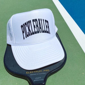 May include: White trucker hat with black text that reads "Pickleballer" on a white mesh back. The hat is sitting on top of a black pickleball paddle with the text "Electrum Pro Designed in California USAPA Approved".