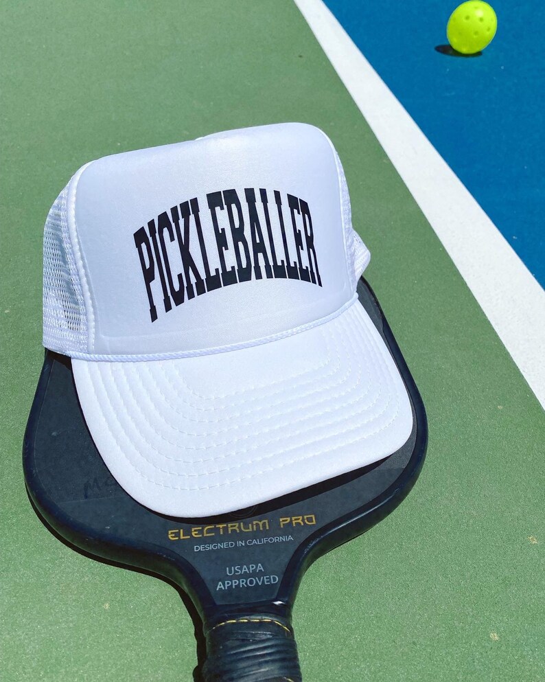 May include: White trucker hat with black text that reads "Pickleballer" on a white mesh back. The hat is sitting on top of a black pickleball paddle with the text "Electrum Pro Designed in California USAPA Approved".