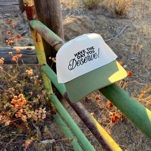 以下が含まれることがあります： 黒で刺繍された「Have the day you deserve!」という文字が入った白と緑の野球帽