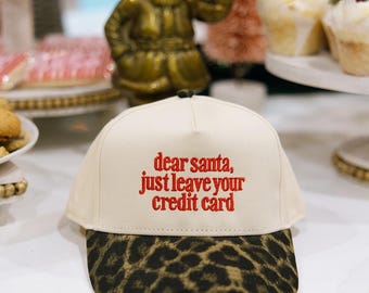 May include: A cream-colored baseball cap with a leopard print brim. The cap features red embroidered text that reads "dear santa, just leave your credit card". The cap is a festive accessory.