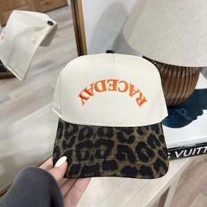Pode incluir: Boné de beisebol branco com a palavra "RACEDAY" bordada em laranja. A aba apresenta uma estampa de leopardo marrom e preto. O boné é refletido em um espelho.