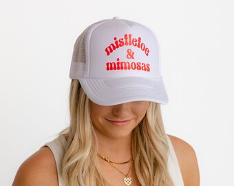 May include: White trucker hat with a mesh back and a curved brim. The hat features the words "mistletoe & mimosas" in red, retro-style lettering across the front. The hat is a casual accessory.