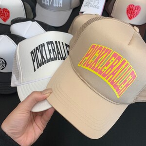 May include: A collection of five trucker hats with different designs. The hats are all black and white with a mesh back. The designs include "Mama", "All You Need Is Love", and "Pickleball".