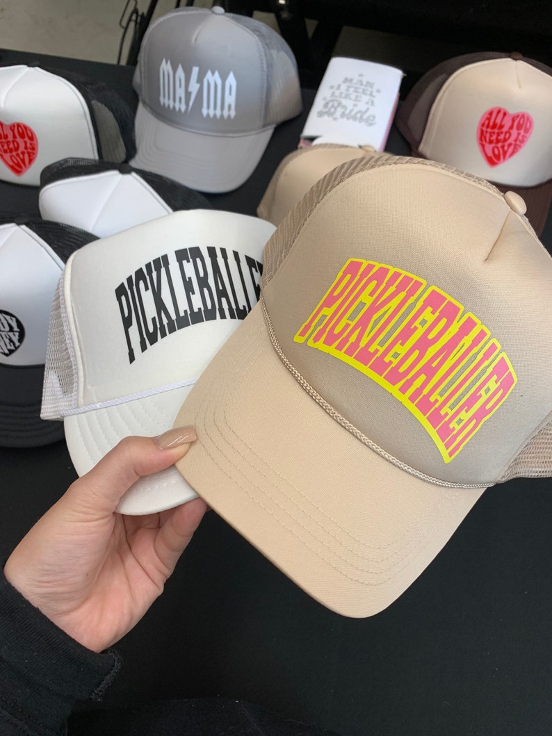 May include: A collection of five trucker hats with different designs. The hats are all black and white with a mesh back. The designs include "Mama", "All You Need Is Love", and "Pickleball".