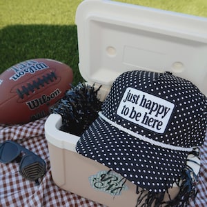 May include: A black baseball cap with white polka dots and a white rope trim across the brim. A white rectangular patch on the front features the black text "just happy to be here". The cap rests on a beige cooler next to a football and sunglasses.