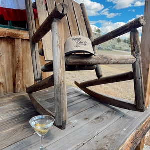 May include: A wooden rocking chair with a tan baseball cap on the seat. The cap has the text "mini fit country" on it. A martini glass with olives sits on the wooden floor in front of the chair.