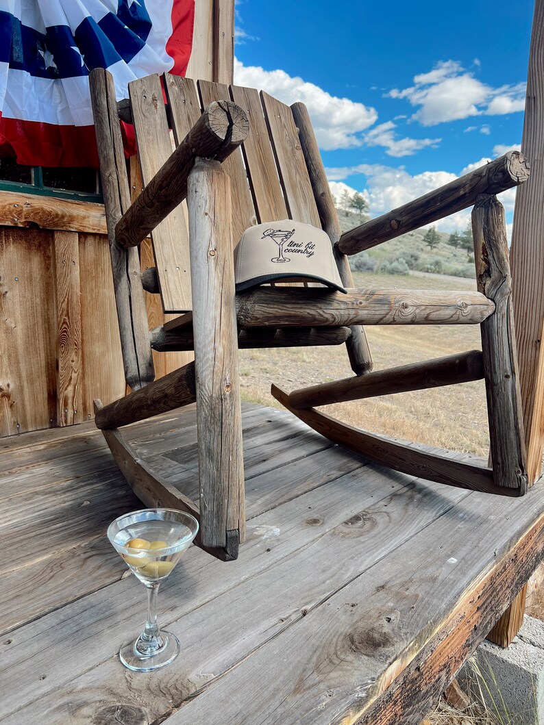 May include: A wooden rocking chair with a tan baseball cap on the seat. The cap has the text "mini fit country" on it. A martini glass with olives sits on the wooden floor in front of the chair.