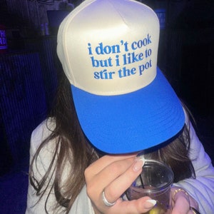 May include: A white and blue baseball cap with the text "i don't cook but i like to stir the pot" embroidered in blue.