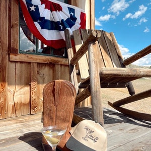 May include: A brown cowboy boot, a martini glass with a green olive, and a tan baseball cap with black trim and the text "mini-file country" on the front. The items are on a wooden porch with a rocking chair and an American flag in the background.