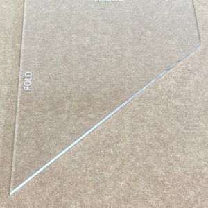 May include: A clear plastic triangle ruler with the word "Medium" printed on the top and the word "FOLD" printed on the left side.