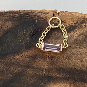 Puede incluir: Un dije de cadena de oro con una piedra rectangular de color púrpura.