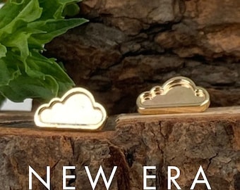 May include: Two gold-colored cloud-shaped stud earrings. The earrings are displayed on a piece of wood with the text "NEW ERA JEWELRY DESIGN" below. Green leaves are visible in the background.