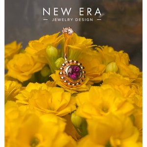 May include: A rose gold belly button ring with a pink gemstone and a clear crystal top. The ring is surrounded by small gold beads.