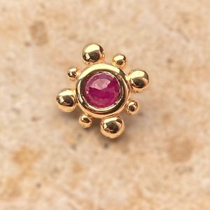 May include: A gold-colored stud earring with a central, round, deep red gemstone. The earring is surrounded by a ring of gold and accented with small, spherical gold embellishments, creating a floral or sunburst design.