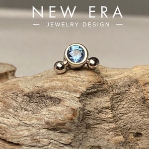 May include: A silver-toned jewelry piece with a central light blue gemstone, flanked by two smaller silver spheres. The jewelry is set against a textured, light brown wooden surface. The text "NEW ERA JEWELRY DESIGN" is displayed above the jewelry.