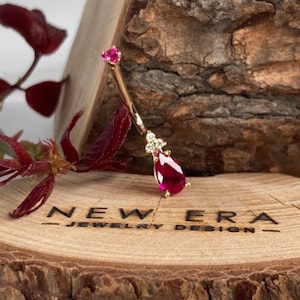 May include: Gold body jewellery with a teardrop-shaped ruby and clear cubic zirconia accents. The jewellery is displayed on a wooden surface with the text "NEW ERA - JEWELRY DESIGN -" engraved on it.