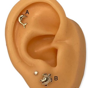 May include: Close-up of a person's ear with two gold earrings. The top earring is a dolphin design and the bottom earring is a turtle design with a small clear stone next to it.