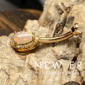 May include: Gold belly button ring with a pear-shaped opal gemstone surrounded by a halo of smaller clear gemstones. The ring is on a wooden surface.