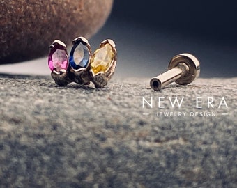 May include: A gold body jewellery piece with three marquise-shaped gemstones. The gemstones are pink, blue, and yellow. The jewellery piece is on a grey textured surface. NEW ERA - JEWELLERY DESIGN - is printed on the surface.
