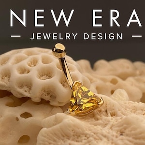 May include: Gold belly button ring with a triangular shaped gemstone. The gemstone is a bright yellow color. The ring is on a white coral-like surface.