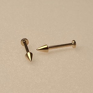 May include: Two gold-coloured body piercing studs with pointed ends. The studs are made of metal and have a smooth finish.