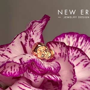 May include: A gold-colored skull-shaped ring rests on a pink and white flower petal. The ring is detailed with facial features. The text "NEW ERA JEWELRY DESIGN" is displayed in white. The background is a soft gray.