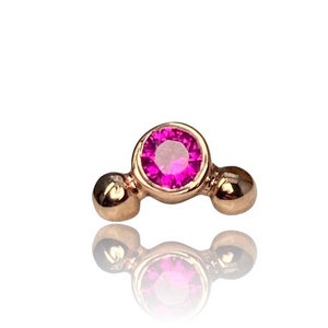 Könnte beinhalten: Ein kleiner, goldfarbener Ohrstecker mit einem leuchtend rosa Edelstein in der Mitte. Der Ohrring hat ein kreisförmiges Design mit einem zentralen Edelstein und zwei kleineren goldenen Kugeln auf beiden Seiten. Der Ohrring spiegelt sich in der darunter liegenden Oberfläche.