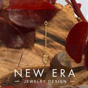 May include: A gold chain with a small gold feather charm hanging from it. The chain is hanging from a piece of wood with red leaves. The text "NEW ERA - JEWELRY DESIGN -" is at the bottom of the image.