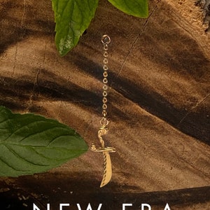 May include: A gold chain necklace with a sword pendant. The pendant is detailed with a curved blade and a twisted handle. The necklace is hanging against a wooden background.