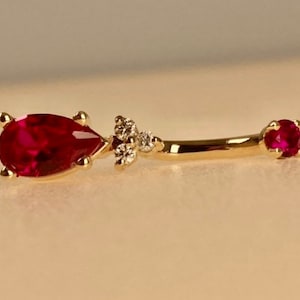 May include: A gold body jewelry piece with a teardrop-shaped ruby gemstone and a smaller round ruby gemstone. The teardrop ruby is set in a prong setting and the round ruby is set in a bezel setting.