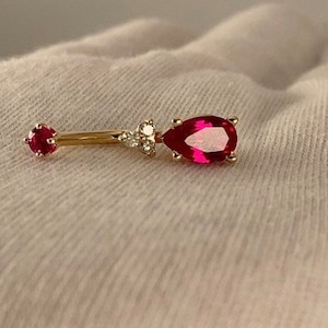 May include: A gold body piercing with a teardrop-shaped ruby gemstone and small clear gemstones on the side.