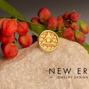 May include: Gold-colored all-seeing eye charm with a triangular design. The charm is on a white rock with red flower buds in the background. The text "NEW ERA - JEWELRY DESIGN -" is visible below the charm.