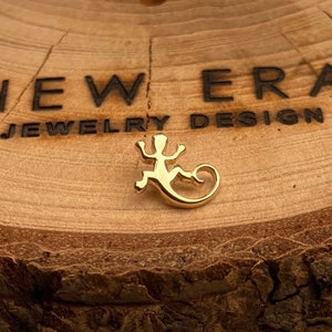 May include: A gold-coloured lizard charm on a wooden surface. The surface has the text "NEW ERA - JEWELRY DESIGN -" engraved on it.