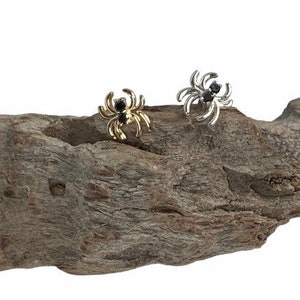 May include: Two spider-shaped earrings, one gold-toned and one silver-toned, each with a black gem at the center. The earrings are displayed on a piece of weathered wood against a white background. The spider earrings are a unique accessory.