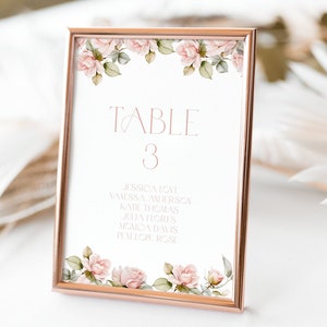 May include: A white table number card with a rose gold frame and floral border. The card reads "TABLE 3" and lists a group of names.