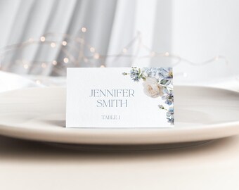Può includere: Un segnaposto rettangolare bianco con il nome stampato JENNIFER SMITH e TABLE 1 in testo grigio scuro. Il lato destro del cartoncino è decorato con un'illustrazione ad acquerello di fiori color azzurro e crema.