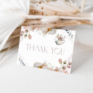 May include: A white card with the words "THANK YOU" in a delicate font, surrounded by watercolor floral illustrations in soft pinks, creams, and blues. A small teapot illustration is also present. The card is set against a blurred background of dried flowers.