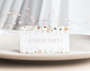 May include: A rectangular white place card featuring a delicate watercolor floral border in shades of pink, green, and yellow, with the name JENNIFER SMITH printed in a thin, centered, uppercase serif font across the middle of the card.