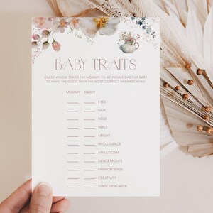 May include: A baby shower game card with the text "BABY TRAITS" at the top, featuring floral illustrations. The card includes prompts for guests to guess traits like eyes, hair, and height. The card is held by a hand.